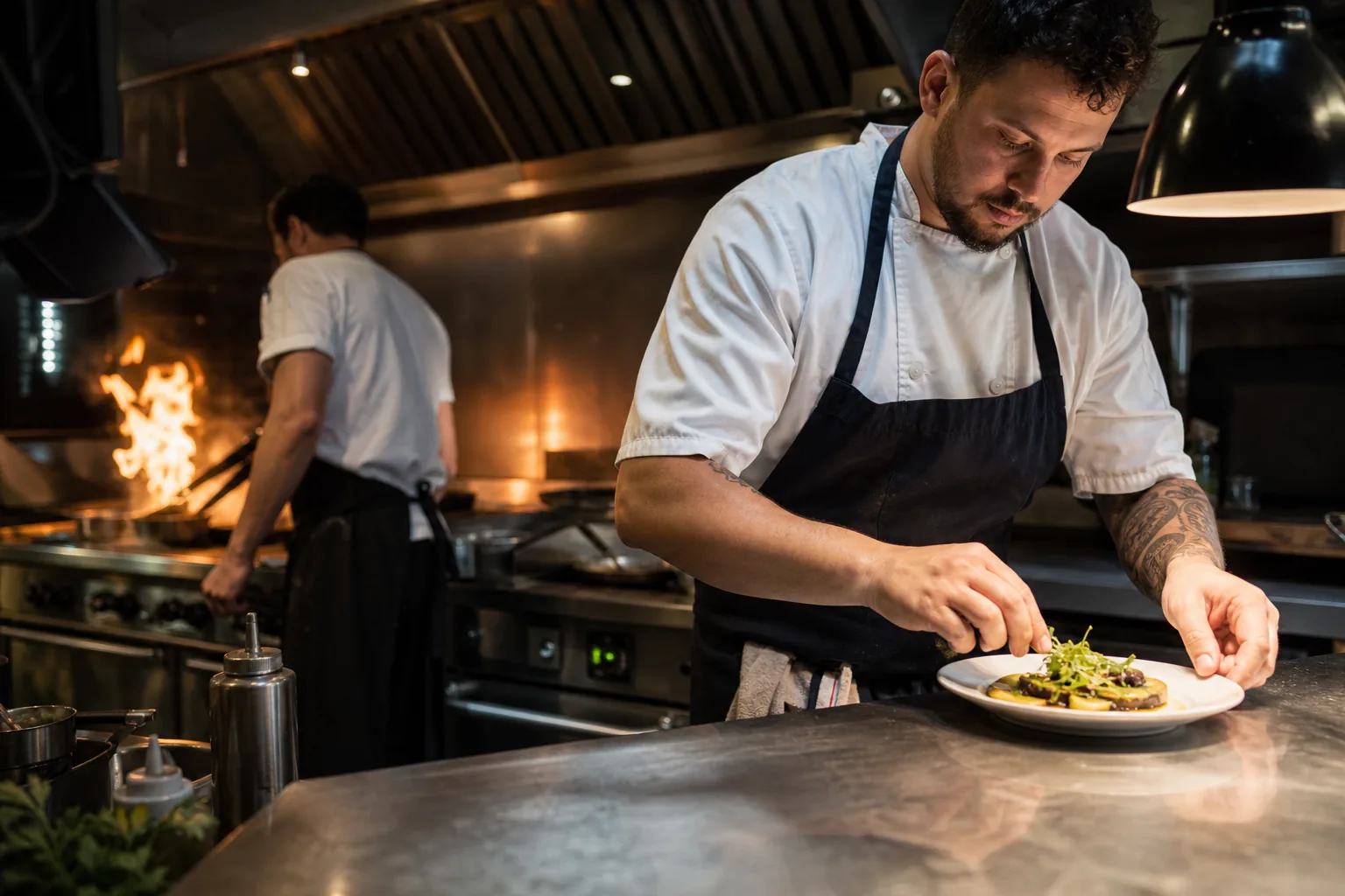 Line cook plating an entree at the pass of a busy restaurant kitchen during dinner service